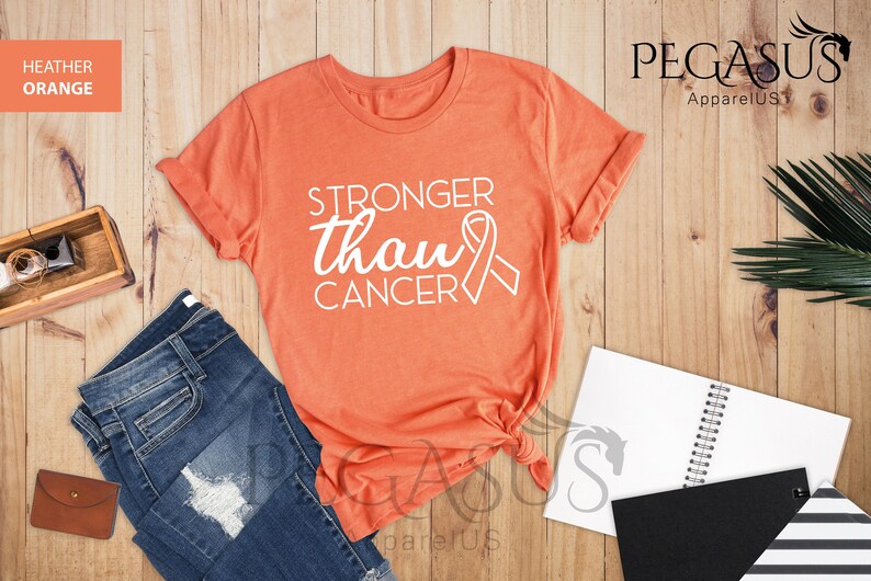 May include: Heather orange t-shirt with the text "Stronger than Cancer" in white with a white ribbon graphic.  The shirt is laid flat on a wood surface.