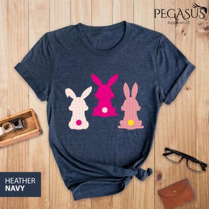 May include: Heather navy t-shirt featuring three Easter bunny designs in pink and white. The bunnies have patterned bodies and white or yellow tail accents. The shirt has the text "PEGASUS ApparelUS" and "HEATHER NAVY".