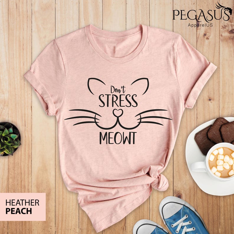 Op de afbeelding: Een roze T-shirt met een zwarte kattenkop-afbeelding en de tekst "Don't Stress Meowt".
