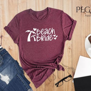 Pu&ograve; includere: T-shirt color tortora con il testo "Beach Bride" in lettere bianche, con palme e cuori.