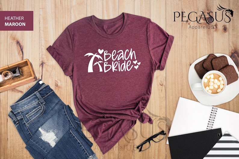 Pu&ograve; includere: T-shirt color tortora con il testo "Beach Bride" in lettere bianche, con palme e cuori.
