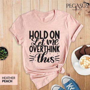 May include: A pink t-shirt with the text "Hold on Let me overthink this" in black lettering. The shirt is on a wooden surface with a cup of coffee and a plate of cookies.