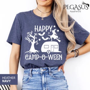 May include: Heather navy blue t-shirt with a white graphic that says "HAPPY CAMP-O-WEEN". The design includes a tree, bats, a witch on a broom, a camper, and stars. The shirt is a crew neck.