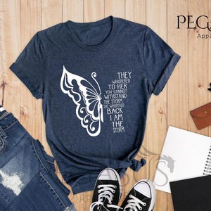 Op de afbeelding: Een marineblauw t-shirt met een witte vlinderprint en de tekst "They whispered to her you cannot withstand the storm she whispered back I am the storm".