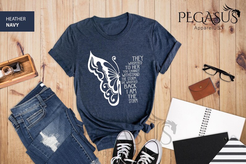 Op de afbeelding: Een marineblauw t-shirt met een witte vlinderprint en de tekst "They whispered to her you cannot withstand the storm she whispered back I am the storm".