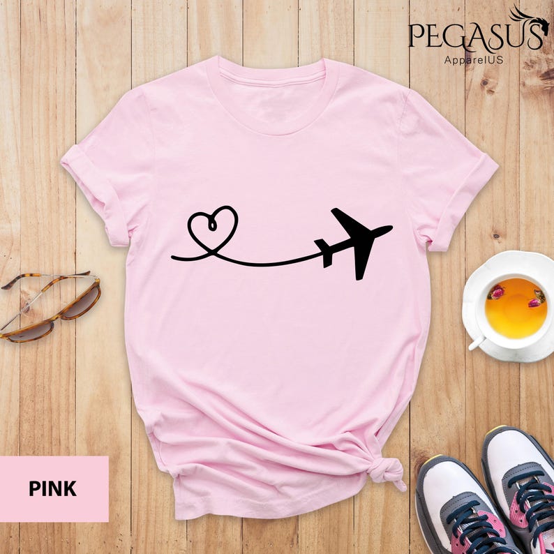 Puede incluir: Camiseta rosa con un avi&oacute;n y un coraz&oacute;n negros. El gr&aacute;fico muestra un coraz&oacute;n con una l&iacute;nea que se extiende hasta un avi&oacute;n. El texto "PEGASUS ApparelUS" est&aacute; en la esquina superior derecha de la imagen.