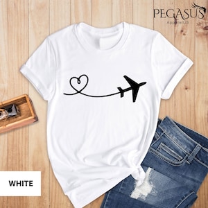 Puede incluir: Camiseta blanca con un gr&aacute;fico de avi&oacute;n y coraz&oacute;n negro. El gr&aacute;fico muestra un coraz&oacute;n con una l&iacute;nea que se extiende a un avi&oacute;n. El texto "PEGASUS ApparelUS" est&aacute; en la esquina superior derecha de la imagen.