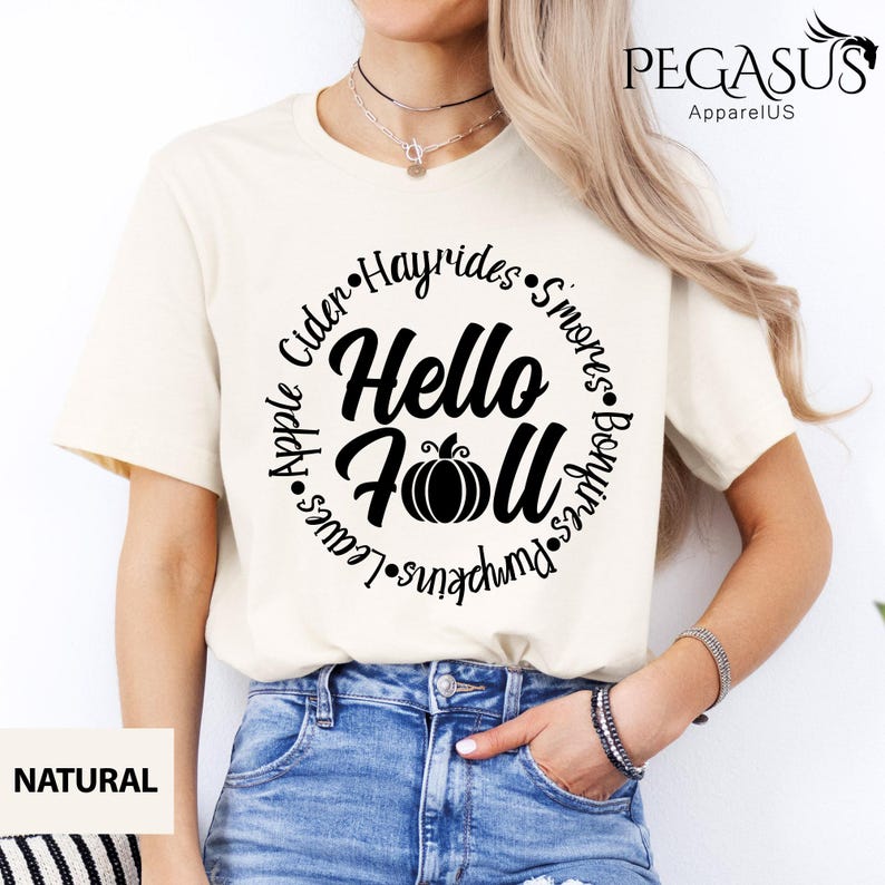 May include: Cream-colored t-shirt with a round black graphic that says "Hello Fall" with a pumpkin illustration. The words "Apple", "Cider", "Hayrides", "S'mores", "Bonfires", "Pumpkins", and "Leaves" are in a circle around the main text.