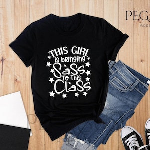 K&ouml;nnte beinhalten: Schwarzes T-Shirt mit wei&szlig;em Text, der "This girl is bringing sass to this class" lautet, mit wei&szlig;en Sternen um den Text herum.