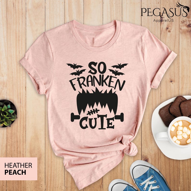 Pu&ograve; includere: T-shirt color pesca con grafica nera che dice "So Franken Cute" con pipistrelli e un design di mostro di Frankenstein. La maglietta &egrave; piegata su una superficie di legno con biscotti e cioccolata calda.