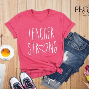 May include: A pink t-shirt with the text "TEACHER STRONG" in white with a white heart in the middle of the word "STRONG".