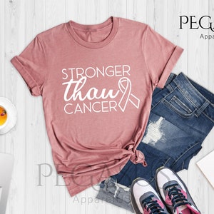 May include: A heather mauve t-shirt with the text "Stronger than Cancer" and a pink ribbon graphic. The shirt is laid out on a white wooden surface with a pair of jeans, a notebook, and a cup of tea.