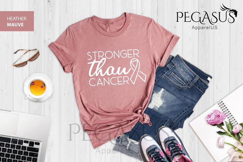 May include: A heather mauve t-shirt with the text "Stronger than Cancer" and a pink ribbon graphic. The shirt is laid out on a white wooden surface with a pair of jeans, a notebook, and a cup of tea.
