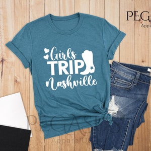 May include: A deep teal t-shirt with the text "Girls Trip Nashville" in white, featuring hearts and a cowboy boot graphic. The shirt is paired with blue jeans on a wooden surface.