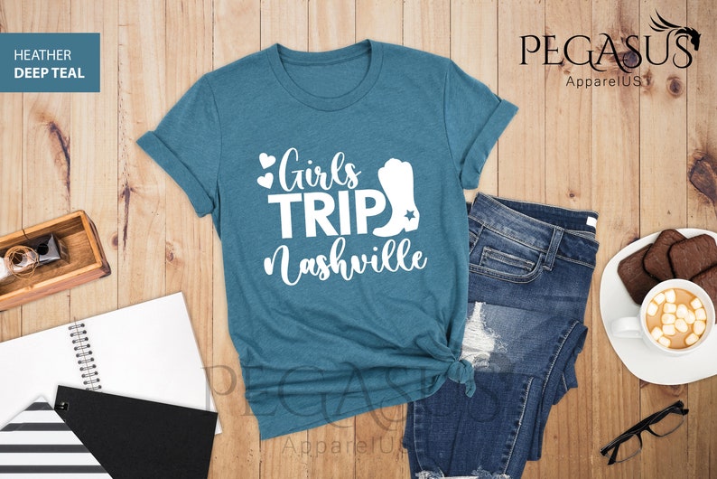 May include: A deep teal t-shirt with the text "Girls Trip Nashville" in white, featuring hearts and a cowboy boot graphic. The shirt is paired with blue jeans on a wooden surface.