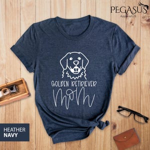 Op de afbeelding: Een heideblauw t-shirt met een witte omtrek van een golden retriever hond en de tekst "Golden Retriever Mom" in wit.