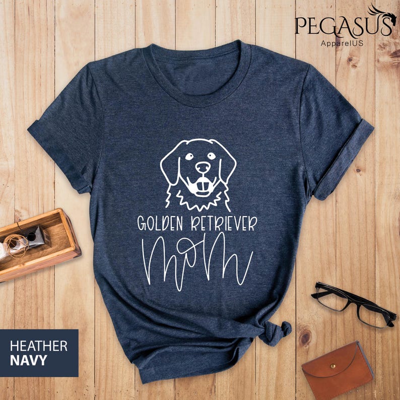 Op de afbeelding: Een heideblauw t-shirt met een witte omtrek van een golden retriever hond en de tekst "Golden Retriever Mom" in wit.