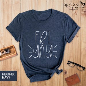 May include: Heather navy blue t-shirt with the word "FRIYAY" in white script. The shirt is displayed on a wooden surface with accessories like glasses and a small leather wallet.