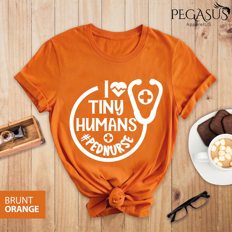 Op de afbeelding: Oranje T-shirt met een witte afbeelding met de tekst "I love tiny humans #pednurse" met een stethoscoop en een hartontwerp.