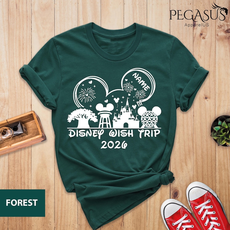 May include: Forest green t-shirt with a white graphic featuring Mickey Mouse ears, fireworks, a castle, and the text "Disney Wish Trip 2026." The word "NAME" is in a bubble. The brand name "Pegasus ApparelUS" is at the top.