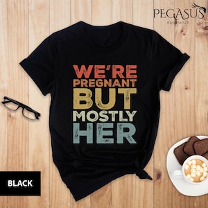 May include: Black t-shirt with a vintage-style graphic that says "We're Pregnant But Mostly Her".