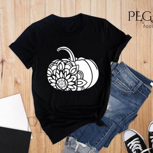 May include: Black t-shirt featuring a white graphic of a pumpkin with a sunflower design. The t-shirt is lying on a wooden surface with a pair of jeans, a notebook, and a cup of coffee.