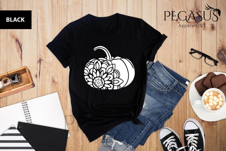 May include: Black t-shirt featuring a white graphic of a pumpkin with a sunflower design. The t-shirt is lying on a wooden surface with a pair of jeans, a notebook, and a cup of coffee.