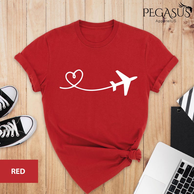 Puede incluir: Camiseta roja con un avi&oacute;n y un coraz&oacute;n blancos. El avi&oacute;n est&aacute; volando lejos de un coraz&oacute;n. El texto "PEGASUS ApparelUS" est&aacute; en la esquina superior derecha de la imagen.