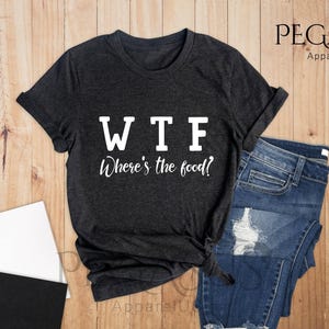 May include: A dark heather grey t-shirt with the text "WTF Where's the food?" printed in white on the front. The shirt is laid flat on a wooden surface with a pair of jeans and a cup of coffee.