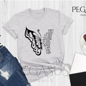 Op de afbeelding: Een grijs t-shirt met een zwarte vlinderprint en de tekst "They whispered to her you cannot withstand the storm she whispered back I am the storm".