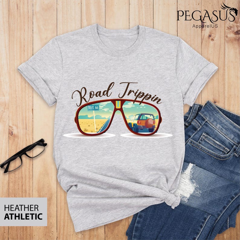 Peut inclure: Un t-shirt gris chin&eacute; avec le texte "Road Trippin" et une paire de lunettes de soleil avec une route et une voiture refl&eacute;t&eacute;es dans les verres. Les lunettes ont une monture marron et des verres jaunes.
