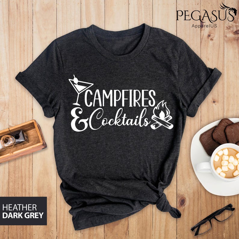 May include: Dark heather gray t-shirt with the text "Campfires & Cocktails" in white. The design includes a cocktail glass and a campfire graphic. The shirt is displayed on a wooden surface with a cup of hot chocolate and glasses.