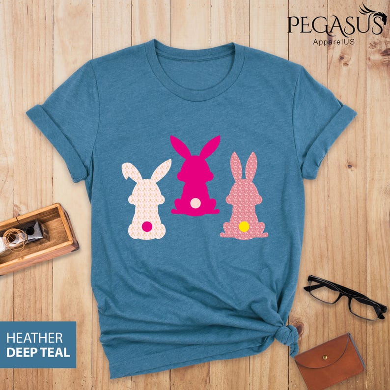 May include: A teal-colored t-shirt featuring three bunny silhouettes in pink and white, each with a contrasting dot. The shirt has rolled-up sleeves and the text "HEATHER DEEP TEAL" is visible. The shirt is on a wooden surface.