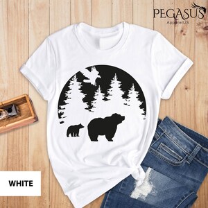 Puede incluir: Camiseta blanca con un dise&ntilde;o de silueta negra de un oso y un cachorro en un bosque con un p&aacute;jaro volando por encima. El dise&ntilde;o est&aacute; dentro de un c&iacute;rculo.