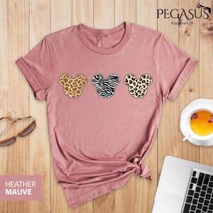 May include: A heather mauve t-shirt with a graphic of three Mickey Mouse heads. Each head is a different animal print: giraffe, zebra, and leopard.