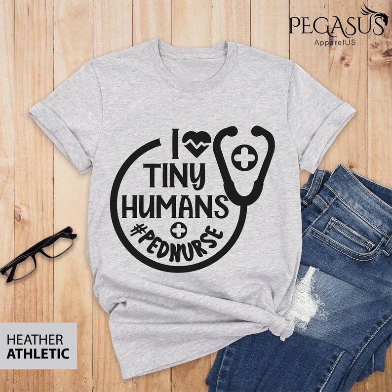 Op de afbeelding: Een heidegrijs T-shirt met een zwarte afbeelding waarop staat "I love tiny humans #pednurse" met een stethoscoop en een hartontwerp.