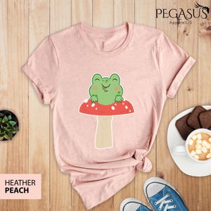 May include: A pink t-shirt with a green frog sitting on a red and white mushroom. The frog is smiling and has its tongue sticking out.