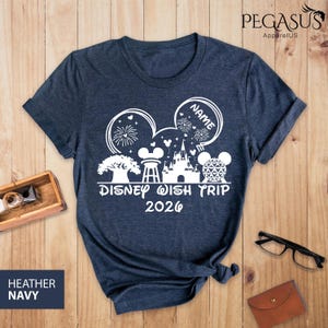 May include: Heather navy blue t-shirt with a white graphic design. The design includes Mickey Mouse ears, a castle, and the text "Disney Wish Trip 2026". The shirt also has the word "Name" in a circle.