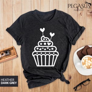 May include: A dark heather grey t-shirt with a white cupcake graphic. The cupcake has white frosting, sprinkles, and hearts.