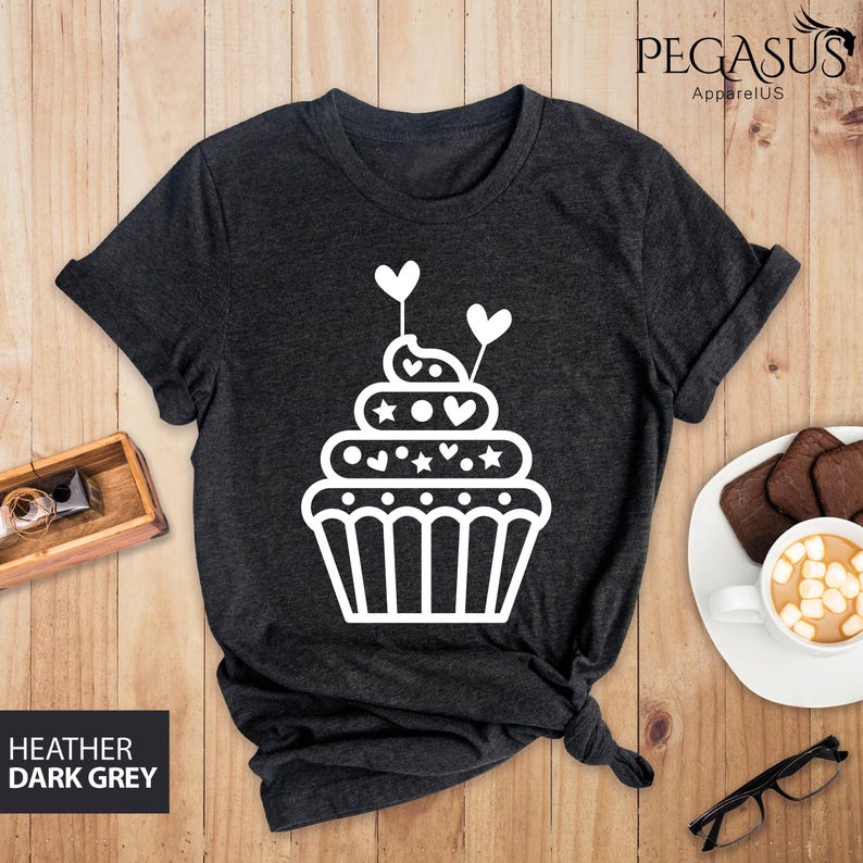 May include: A dark heather grey t-shirt with a white cupcake graphic. The cupcake has white frosting, sprinkles, and hearts.
