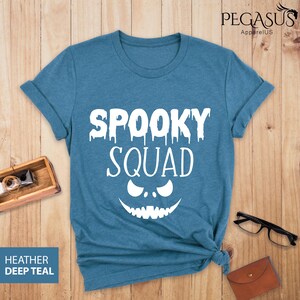 May include: A teal t-shirt with the words "SPOOKY SQUAD" in white, featuring a dripping font and a jack-o'-lantern face. The shirt is made of a soft material and is perfect for Halloween.