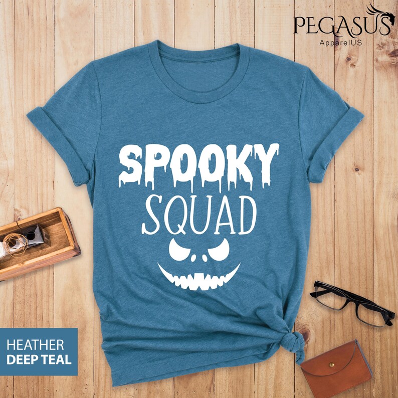 May include: A teal t-shirt with the words "SPOOKY SQUAD" in white, featuring a dripping font and a jack-o'-lantern face. The shirt is made of a soft material and is perfect for Halloween.