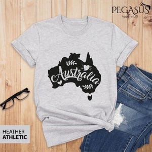 Op de afbeelding: Een heidegrijs T-shirt met een zwarte omtrek van het continent Australi&euml; en het woord "Australia" in een gestileerd lettertype. Het ontwerp bevat kleine hartjes en bladeren.