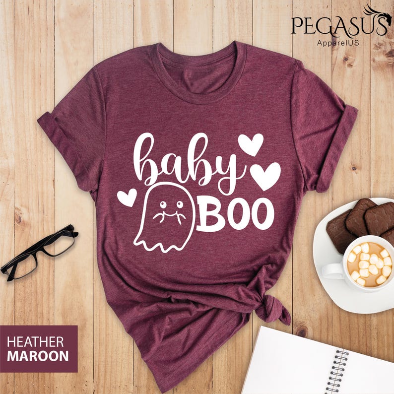 May include: Heather maroon t-shirt with the text "baby BOO" in white, featuring a ghost and hearts. The shirt is styled on a wooden surface with a pair of glasses, a notepad, and a cup of hot chocolate.