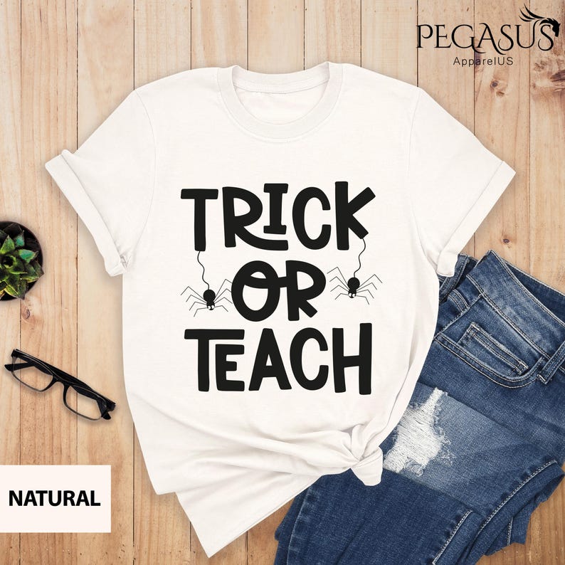 May include: White t-shirt with the text "TRICK OR TEACH" in black, featuring spider graphics. The shirt is paired with blue jeans and rests on a wooden surface. The brand name "PEGASUS ApparelUS" is visible.