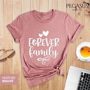 Pode incluir: Uma camiseta de cor malva com o texto "FOREVER family" em letras brancas com dois pequenos cora&ccedil;&otilde;es acima do texto. A camiseta &eacute; feita de um material macio e confort&aacute;vel e &eacute; perfeita para o uso di&aacute;rio.