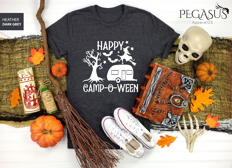 May include: Dark grey t-shirt with a white graphic reading "HAPPY CAMP-O-WEEN", featuring a tree, witch, bats, and a camper. The image includes Halloween decorations such as pumpkins, a broom, and a skull.