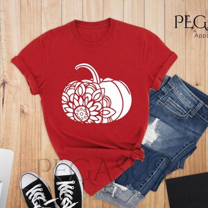 May include: Red t-shirt featuring a white graphic of a pumpkin with a mandala design. The t-shirt is displayed on a wooden surface with a pair of jeans and a laptop.