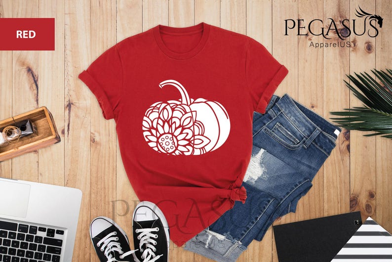 May include: Red t-shirt featuring a white graphic of a pumpkin with a mandala design. The t-shirt is displayed on a wooden surface with a pair of jeans and a laptop.