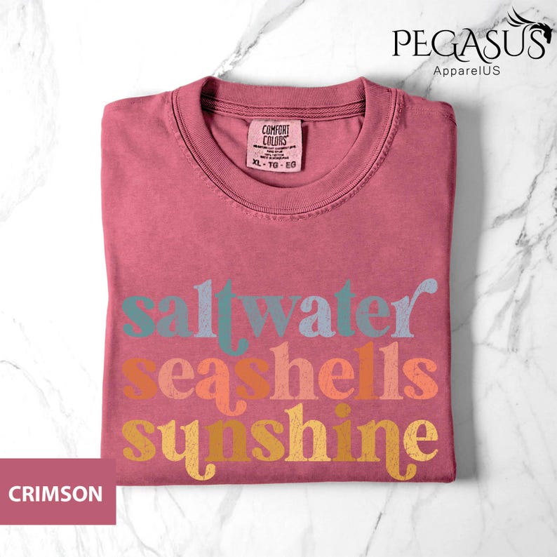 May include: Crimson-colored t-shirt with the words "saltwater seashells sunshine" in colorful, retro-style lettering. The shirt has a crew neck and a tag that reads "Comfort Colors". The Pegasus ApparelUS logo is in the upper right corner.